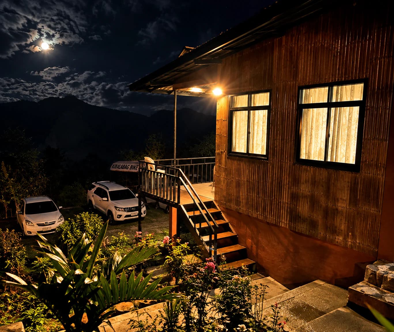 The Family Cottage at night, its windows lit warm under a full moon, with steps up to the veranda